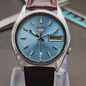 May include: A Seiko 5 Sports automatic wristwatch with a light blue sunray dial, silver-tone hour markers, and a day-date window at the three o'clock position. The watch features a stainless steel case and a brown leather strap with visible stitching.