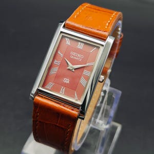 Könnte beinhalten: Eine rechteckige Seiko Quartz Armbanduhr mit silberfarbenem Gehäuse, einem strukturierten terrakottafarbenen Zifferblatt mit römischen Ziffern sowie einem leuchtend orangefarbenen Lederarmband mit Krokodilprägung und Nähten.