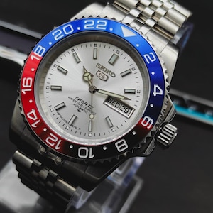 May include: A stainless steel Seiko 5 Sports automatic watch with a silver dial, luminous hands, and a day-date window. The bezel features a red and blue insert with white numerals. The watch includes a silver-tone metal link bracelet.