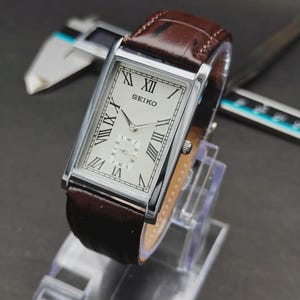 May include: A rectangular Seiko wristwatch with a silver-tone case, a white dial featuring black Roman numeral hour markers, and a brown leather strap with a textured finish. The watch includes a small sub-dial for seconds at the six o'clock position.