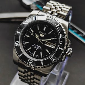 Op de afbeelding: Een zilverkleurig Seiko 5 Sports automatisch polshorloge met een zwarte wijzerplaat, een draaibare zwarte ring met witte cijfers en een roestvrijstalen horlogeband in jubilee-stijl. De wijzerplaat heeft lichtgevende wijzers en een dag-datumvenster.