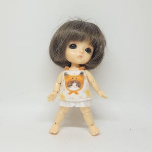 May include: A small doll with a brown bob haircut and large black eyes. The doll wears a white dress with a yellow and orange frog design on the front. The doll's skin is a pale peach color.