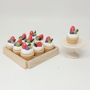 以下が含まれることがあります： 白いフロスティングを施したミニチュアカップケーキ。赤いイチゴ、紫と緑のベリー、チョコレート削りくずがトッピングされています。カップケーキは木製のトレイに並べられており、1つは小さな白い台座にあります。