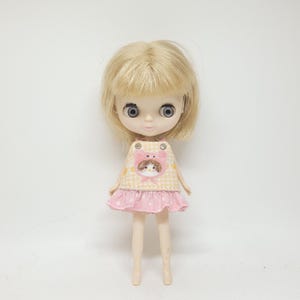 May include: A small doll with a blonde wig and large eyes. The doll wears a pink and yellow dress with a hedgehog graphic on the front. The dress has a pink skirt with white polka dots. The doll is standing on a white surface.