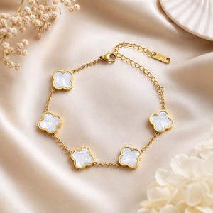 May include: A gold-toned chain bracelet featuring five evenly spaced, four-leaf clover charms with white, iridescent centers. The bracelet includes a delicate link chain, a lobster claw clasp, and a small rectangular tag engraved with S.STEEL.