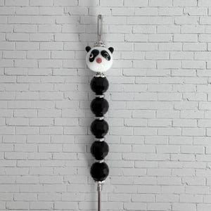 May include: A decorative pen topper featuring a white and black panda bear bead at the top, followed by five faceted black beads separated by small silver-toned metal spacers, all mounted on a thin silver-toned metal rod with a loop at the end.