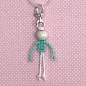 Op de afbeelding: Een kleine hanger in de vorm van een poppetje met een textuurrijke lichtgrijze bolvormige kop, een romp van geknoopt mintgroen koord en twee witte koordbenen. Het figuurtje is bevestigd aan een zilverkleurige metalen karabijnsluiting.