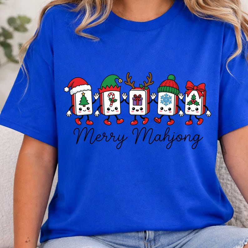Może przedstawiać: Kr&oacute;lewsko-niebieski t-shirt z grafiką przedstawiającą pięć rysunkowych płytek Mahjong trzymających się za ręce. Każda płytka ma inny świąteczny dodatek, w tym czapkę Mikołaja, czapkę elfa, rogi renifera, zimową czapkę i czerwoną kokardę. Poniżej znajduje się napis Merry Mahjong.