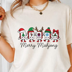 Może przedstawiać: Kremowy t-shirt z grafiką przedstawiającą pięć antropomorficznych płytek mahjonga trzymających się za ręce. Każda płytka ma inny świąteczny dodatek, w tym czapki, poroże i kokardy. Poniżej znajduje się nadruk z napisem Merry Mahjong.