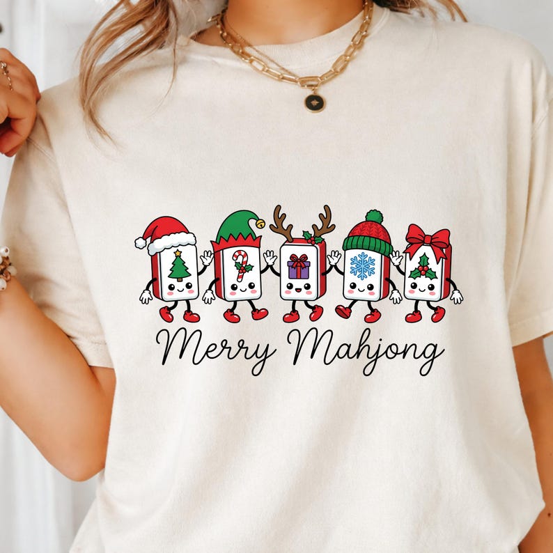 Może przedstawiać: Kremowy t-shirt z grafiką przedstawiającą pięć antropomorficznych płytek mahjonga trzymających się za ręce. Każda płytka ma inny świąteczny dodatek, w tym czapki, poroże i kokardy. Poniżej znajduje się nadruk z napisem Merry Mahjong.