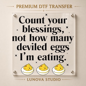 May include: A clear square acrylic sign featuring the black text "Count your blessings, not how many deviled eggs I'm eating." with small star accents and three illustrated deviled eggs at the bottom. The top reads "PREMIUM DTF TRANSFER" and the bottom "LUNOVA STUDIO".