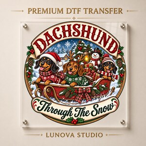 Peut inclure: Un transfert DTF premium représentant trois teckels dans un traîneau rouge au milieu d'une scène hivernale enneigée. Le motif inclut le texte DACHSHUND et Through The Snow, orné de houx, de pommes de pin, d'un bonhomme de neige et de cadeaux.
