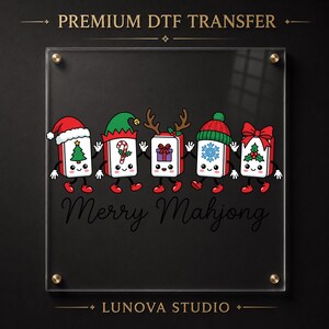 Może przedstawiać: Transfer DTF Premium przedstawiający pięć klock&oacute;w mahjonga w stylu kresk&oacute;wkowym z akcesoriami świątecznymi, takimi jak czapka Mikołaja, czapka elfa, poroże renifera, czapka zimowa i czerwona kokarda. Napis Merry Mahjong oraz Lunova Studio na akrylu.