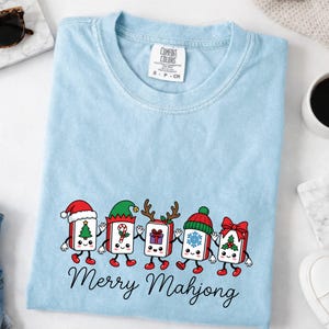 Może przedstawiać: Jasnoniebieski t-shirt z okrągłym dekoltem i grafiką przedstawiającą pięć antropomorficznych płytek mahjonga ubranych w świąteczne akcesoria, takie jak czapki Mikołaja i rogi renifera, z napisem Merry Mahjong wydrukowanym czarną czcionką.
