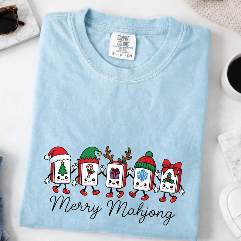 Może przedstawiać: Jasnoniebieski t-shirt z okrągłym dekoltem i grafiką przedstawiającą pięć antropomorficznych płytek mahjonga ubranych w świąteczne akcesoria, takie jak czapki Mikołaja i rogi renifera, z napisem Merry Mahjong wydrukowanym czarną czcionką.