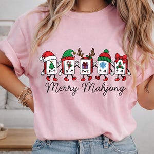 Może przedstawiać: Jasnor&oacute;żowy t-shirt z grafiką przedstawiającą pięć rysunkowych płytek Mahjong trzymających się za ręce. Każda płytka ma inny świąteczny dodatek, w tym czapkę Mikołaja, czapkę elfa, rogi renifera, zimową czapkę i czerwoną kokardę. Poniżej znajduje się napis Merry Mahjong.