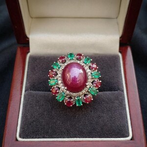 May include: A ring featuring a large oval cabochon ruby centre stone surrounded by a halo of alternating oval-cut rubies and emeralds, accented with small diamonds, presented in a dark wood box with the text ZelmoraJewels printed inside.