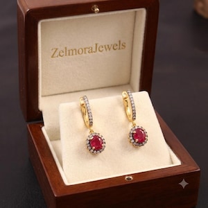 Puede incluir: Un par de pendientes colgantes de oro con rubíes rojos de talla ovalada rodeados de pequeños diamantes blancos. Los pendientes se presentan en un estuche de madera oscura con el texto ZelmoraJewels impreso en dorado en el forro interior crema.