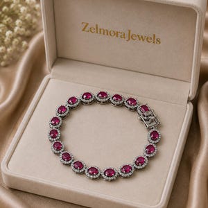 Pode incluir: Uma pulseira de tom prateado com uma série de rubis vermelho-escuros de corte oval, cada um rodeado por um halo de pequenas pedras transparentes. A pulseira tem um fecho metálico seguro e é apresentada numa caixa de joias bege com a inscrição Zelmora Jewels.
