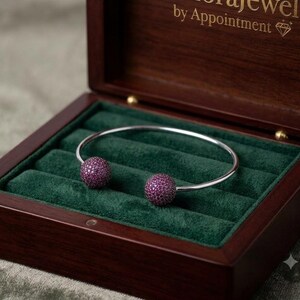 May include: A silver-toned open cuff bracelet featuring two spherical ends encrusted with small, vibrant magenta stones. The bracelet is displayed inside a dark wooden jewellery box with a deep green velvet interior and the text by Appointment.