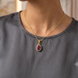 May include: A gold chain necklace featuring an oval-shaped pendant with a deep red ruby center stone surrounded by a halo of small, clear, round-cut white stones. The pendant is suspended from a delicate gold rope chain that rests against the neck.