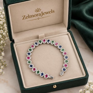 Può includere: Un bracciale color argento con pietre preziose ovali alternate rosse, blu e verdi, ciascuna circondata da un alone di piccole pietre bianche. Il bracciale è esposto all'interno di un portagioie verde scuro con il testo ZelmoraJewels stampato all'interno.