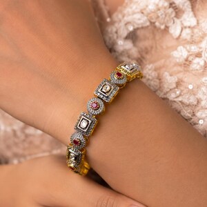 May include: A gold-toned bracelet featuring alternating square and circular links. The square links contain a central clear stone, while the circular links feature a vibrant red stone. The metal surfaces are textured with small, granular details.