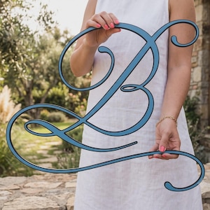 May include: A large, decorative wall hanging in the shape of the cursive letter B. The letter features a smooth, matte blue finish with a thin black border outlining the elegant, flowing script design, creating a bold and clear visual statement.