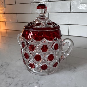 Puede incluir: Un azucarero de cristal transparente y rojo rubí con tapa a juego. El recipiente presenta un patrón geométrico de panal con facetas hexagonales que alternan entre transparente y rojo. Tiene dos asas laterales y un remate facetado en la tapa.