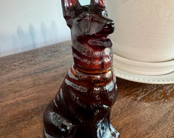 May include: A dark amber-colored glass figurine shaped like a German Shepherd dog in a seated position. The translucent glass sculpture features molded details of the dog's ears, facial features, and fur texture, with a smooth, glossy finish throughout.
