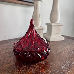 May include: A deep red glass candy dish with a pointed, conical lid. The entire piece features a faceted, diamond-cut pattern across the surface, creating a geometric texture that catches the light. The base is rounded and the lid tapers to a point.
