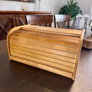 May include: A rectangular wooden bread box featuring a curved, slatted roll-top door. The container is constructed from light-toned wood with a smooth finish, showcasing horizontal slats that form the sliding lid mechanism for the storage unit.