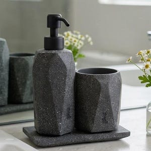 Puede incluir: Un juego de accesorios de baño en color gris oscuro que incluye un dispensador de jabón facetado con bomba negra, un vaso a juego y una bandeja rectangular. Los artículos presentan una textura moteada y están colocados sobre una encimera.
