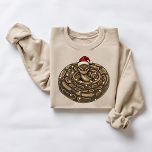 May include: A beige crewneck sweatshirt featuring a graphic of a coiled brown and tan patterned snake wearing a red and white Santa hat. The snake is wrapped in a string of glowing yellow Christmas lights, creating a festive holiday design.