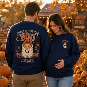 Könnte beinhalten: Ein marineblaues Sweatshirt mit Rundhalsausschnitt, das auf der Rückseite eine große Grafik mit dem Text "OUR LITTLE BOO" und "COMING THIS HALLOWEEN" zeigt, die ein Baby in einer Geisterdecke mit Kürbismütze darstellt, sowie eine kleinere passende Grafik auf der linken Brust.