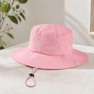May include: A light pink bucket hat featuring an embroidered white bow and the name Sophie in cursive script. The hat includes a wide brim, a chin strap with an adjustable black toggle, and small white metal ventilation eyelets on the side.