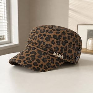 May include: A military-style cap featuring a brown and black leopard print pattern. The hat includes a structured brim, a decorative band around the crown, two dark circular buttons on the side, and the word MAMA in metallic silver lettering.