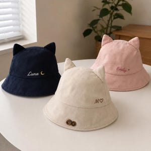 Op de afbeelding: Drie bucket hats met kattenoren aan de bovenkant. De marineblauwe hoed heeft de tekst Luna en een halve maan. De beige hoed heeft de tekst M en een hartje, met twee bruine knopen. De roze hoed heeft de tekst Emily en een klein sterretje.