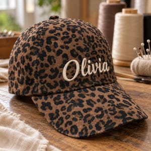 May include: A brown and black leopard print baseball cap featuring the name Olivia embroidered in white script lettering on the front panel. The cap has a structured crown, a curved brim, and a button at the top centre of the headpiece.