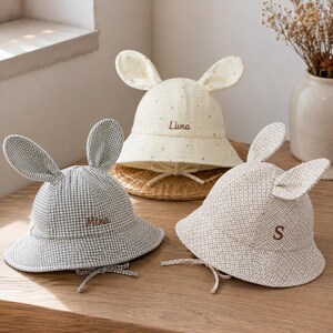 Op de afbeelding: Drie bucket hats met konijnenoren en kinbandjes. Eén is grijs geblokt met de naam Mina, één is crème met gouden stippen en de naam Luna, en één is beige met kleine bruine stippen en de letter S geborduurd op de voorzijde van elke hoed.