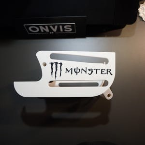 Könnte beinhalten: Eine weiße, 3D-gedruckte Kunststoffhalterung mit dem schwarzen Monster Energy Logo und Markennamen auf der Seite. Das Bauteil hat eine geschwungene, längliche Form mit zwei horizontalen Aussparungen und zwei runden Befestigungslöchern.