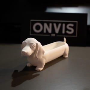 Peut inclure: Un récipient blanc au fini mat en forme de teckel avec un dos creusé pour le rangement. La figurine de chien présente des oreilles tombantes, un long corps et une petite queue. En arrière-plan, un panneau noir rectangulaire affiche le texte ONVIS.