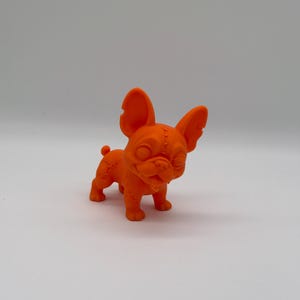 May include: A bright orange figurine of a French Bulldog with large, upright ears and a small tail. The surface of the dog features visible molded stitch patterns across the head, back, and legs, giving it a textured, assembled appearance.