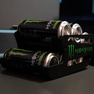 Op de afbeelding: Een zwarte kunststof drankblikjeshouder met meerdere zwarte en groene Monster Energy Zero Sugar blikjes. De zijkant van de houder is voorzien van het groene Monster-logo en de tekst. Het getrapte ontwerp zorgt voor een georganiseerde opslag.