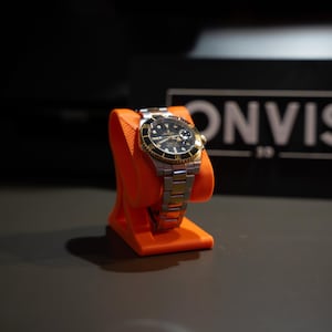 May include: A silver and gold-tone wristwatch with a black dial and rotating bezel, displayed on a bright orange plastic stand. The watch features a metallic link bracelet and a date window. In the background, a black sign displays the text ONVIS.