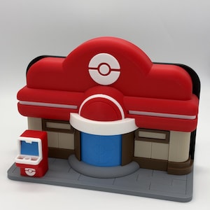 Może przedstawiać: Wydrukowany w 3D model budynku Centrum Pokémon z czerwonym dachem i białym okrągłym emblematem, niebieskimi drzwiami z centralnym logo, beżowymi ścianami i małym czerwonym kioskiem na szarej, wyłożonej kafelkami podstawie. Gładkie, matowe wykończenie.