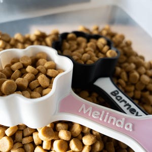 May include: Two paw-shaped pet food scoops filled with brown kibble. One scoop is white with a pink handle featuring the name Melinda in white text. The other scoop is black with a white handle featuring the name Barney in black text.