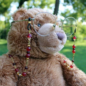 May include: A brown plush teddy bear wearing round gold-rimmed glasses with a decorative beaded chain. The chain features a series of red and dark maroon spherical beads interspersed with small gold-toned metal spacers and links.