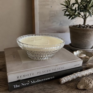 May include: A three-wick candle in a clear, textured glass bowl with a diamond-cut pattern. The candle is placed on top of two stacked books titled Remote Places to Stay and The New Mediterranean, which are resting on a wooden surface.
