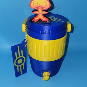 May include: A blue and yellow plastic beverage cooler featuring a yellow band around the center and a yellow spigot at the base. A mushroom cloud charm sits on the lid, and a rectangular blue tag with a yellow circular gear logo hangs from a chain.